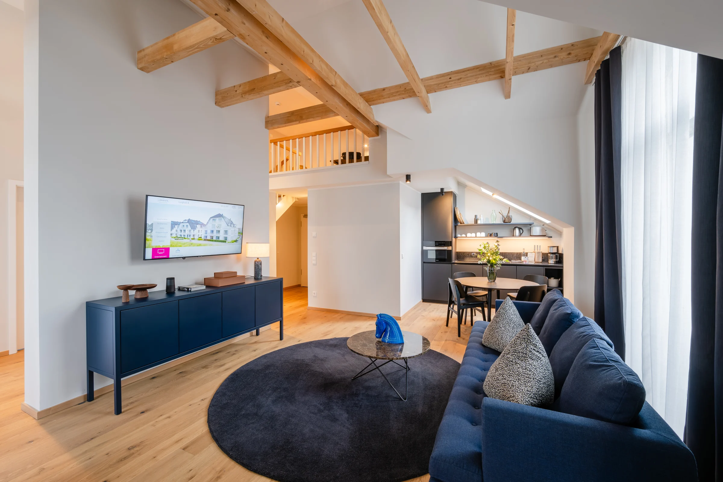 Wohnbereich im LORNSEN-Penthouse mit Holzbalken, dunkelblauem Sofa und offener Küche
